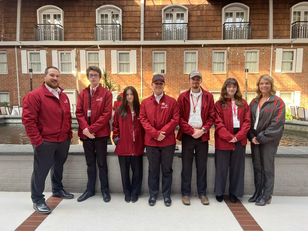 Five students in red jackets stand flanked by two adults at the Desmond Hotel in Albany.