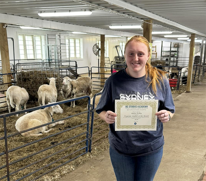 An Ag Academy student holding a certificate in a barn building with sheep penned nearby.