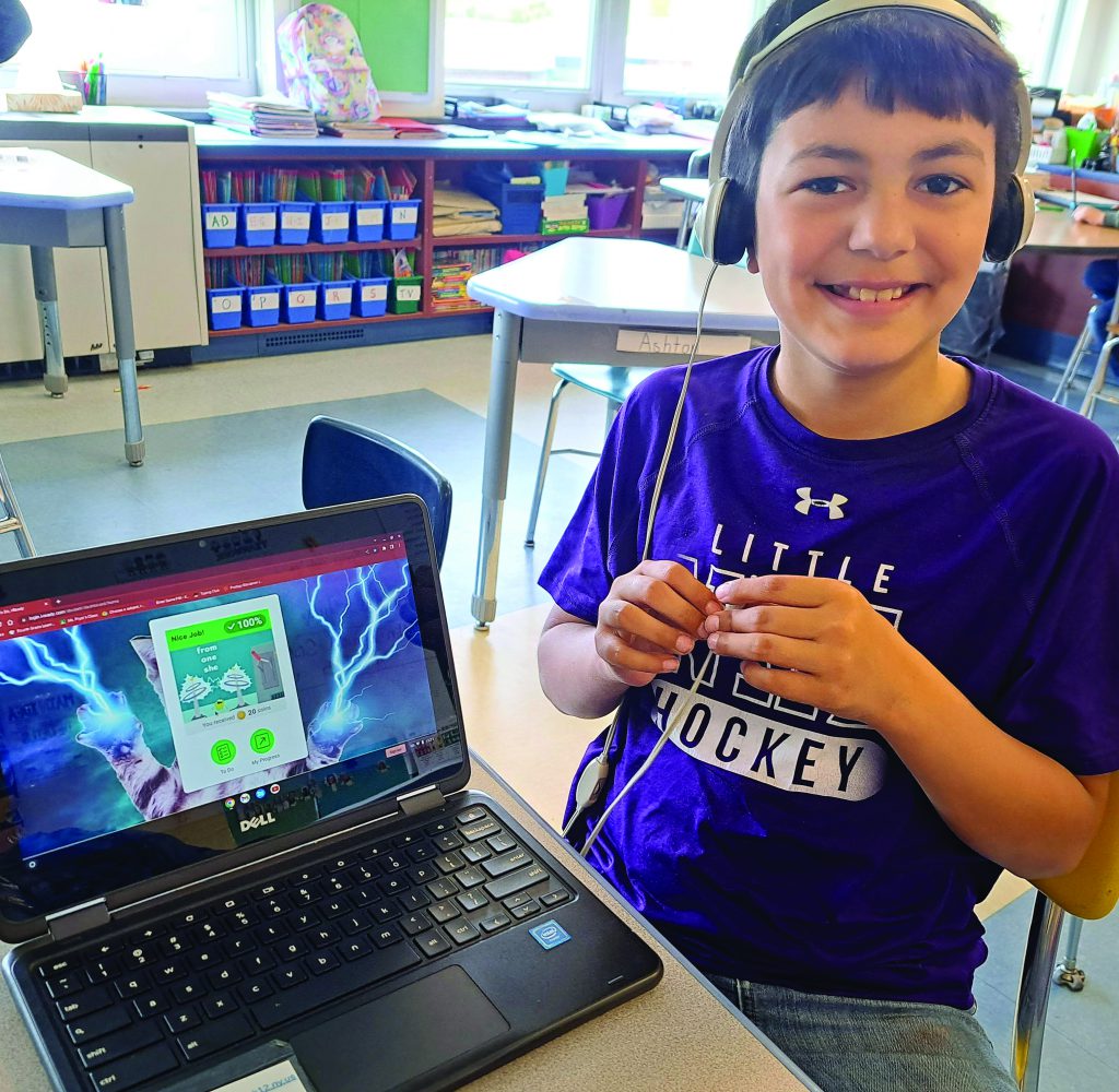 A student wearing headphones sits next to a laptop displaying an educational game.