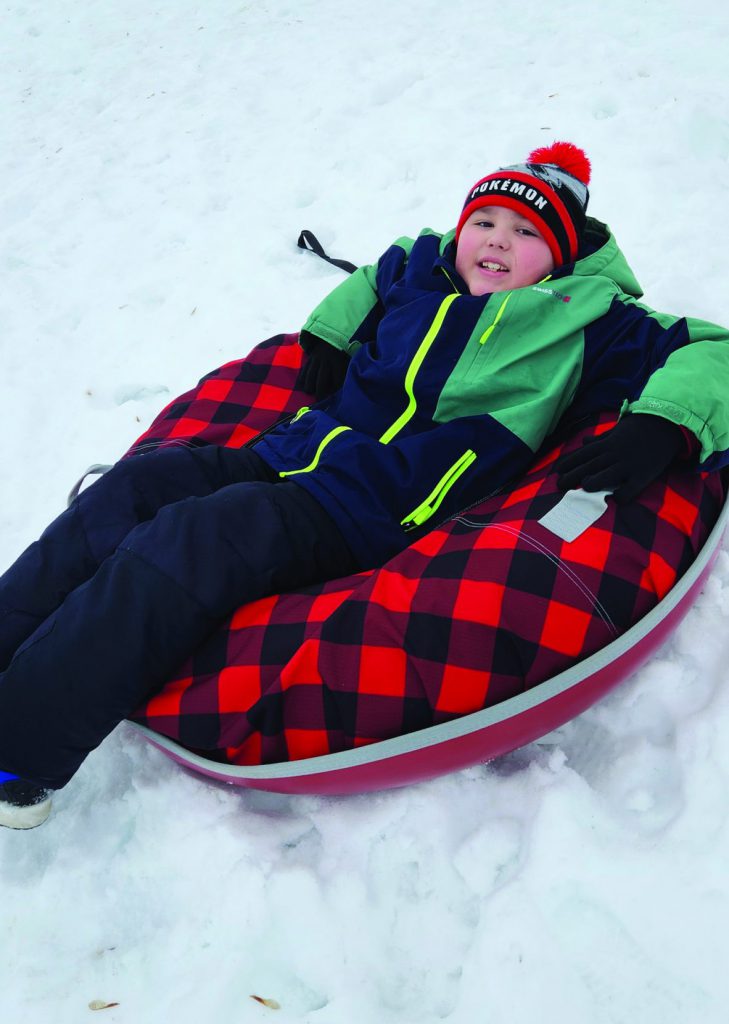 A student bundled in warm weather gear lays on a sled in the snow.