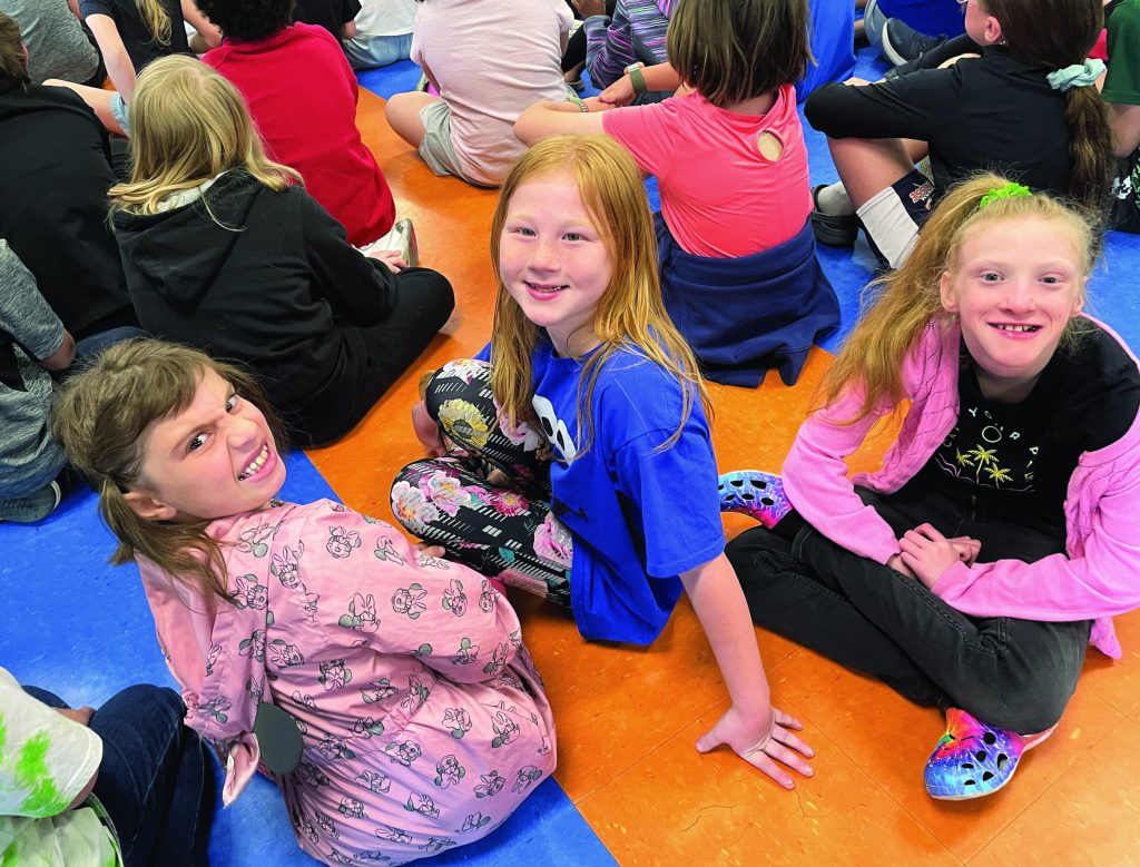 Three young students sitting among other students on the floor turn to face the camera and smile.