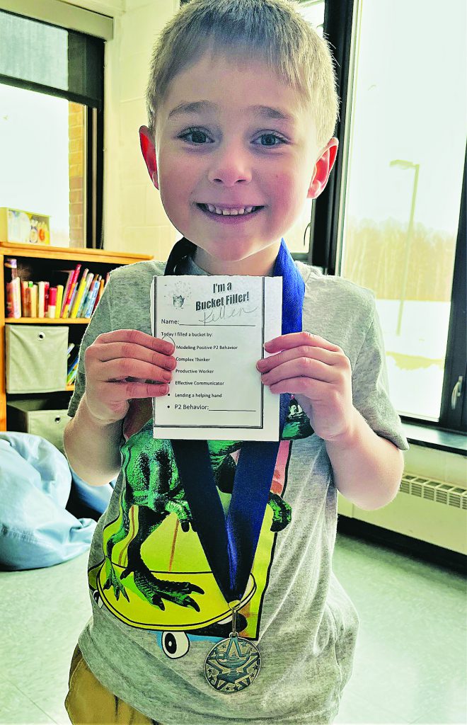 A young child wearing a star medal proudly holds a note indicating that he is a "bucket filler".