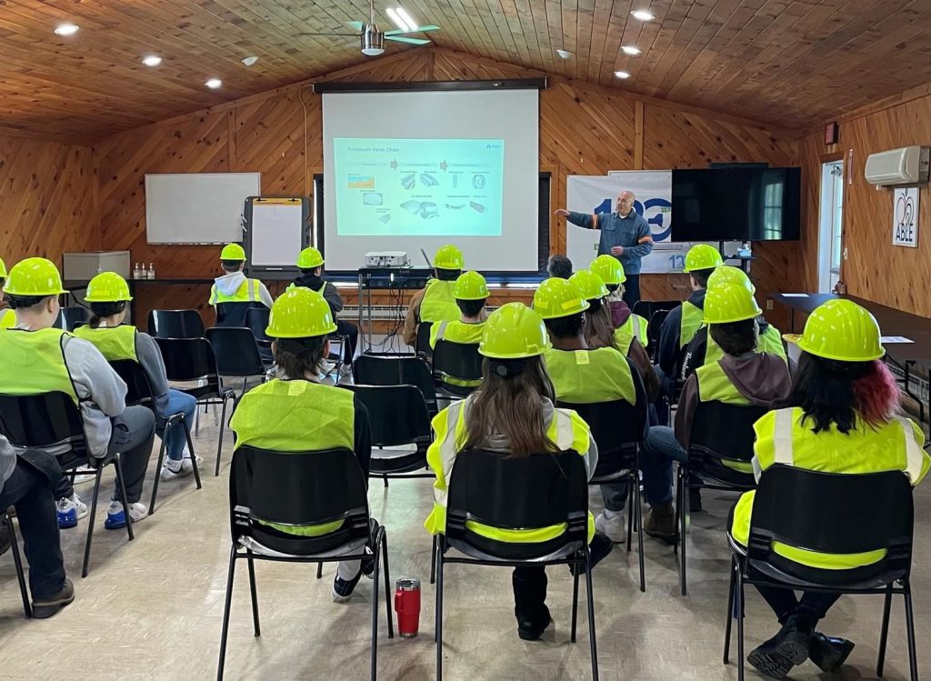 Students are seated in an instruction room as an Alcoa employee speaks to (undetermined) illustrations on a projector screen.