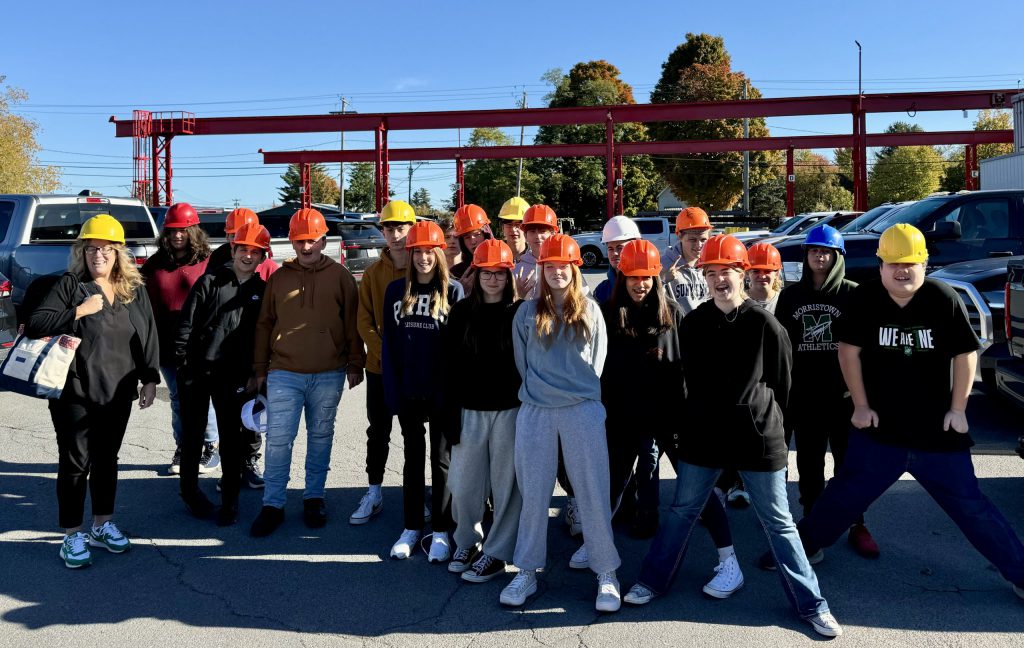 Students in hardhats pose together in front of a steel beam structure in a parking lot.