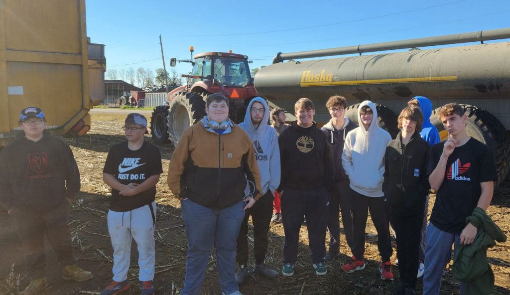 Several students gather outdoors in front of a tractor hitched to a tanker.