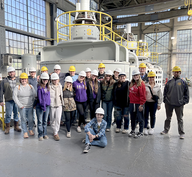 Students wearing hardhats gather in front of very large equipment during their Manufacturing Day tour.