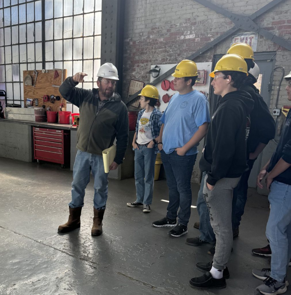 Students wearing hardhats listen to a foreman in a factory setting.