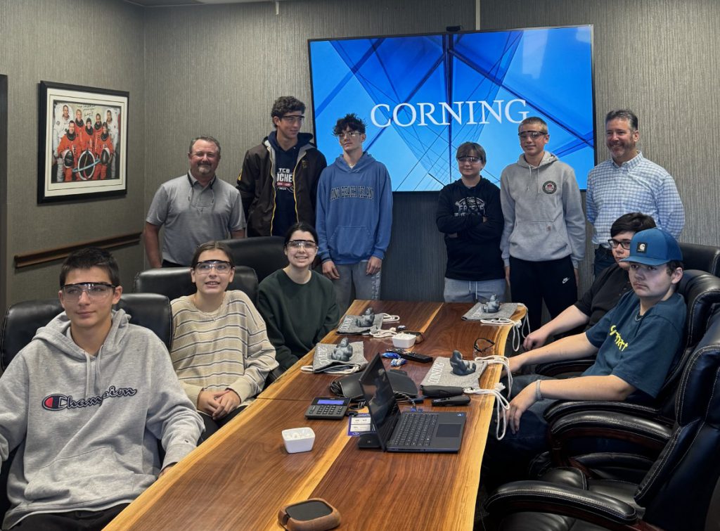 Several students and two business leaders gather around a conference table with a all screen that says "Corning" in the background.