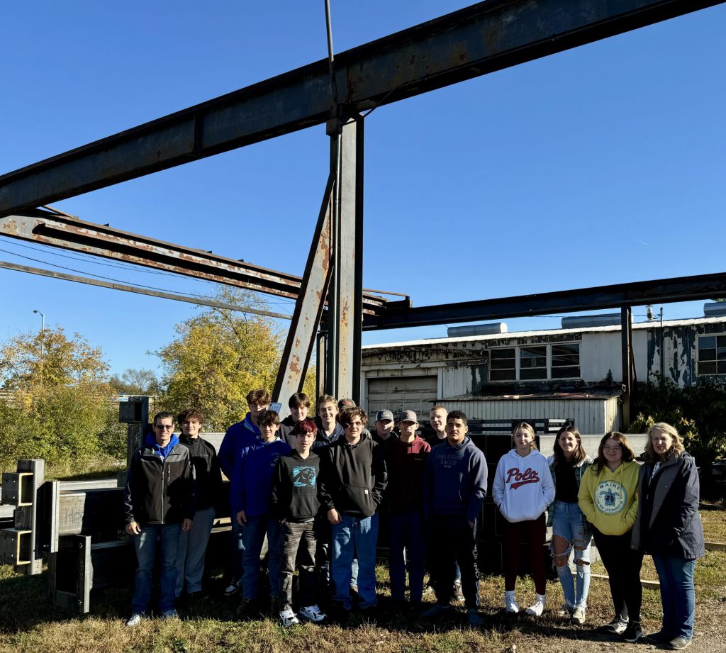 Several students gather together outside underneath a simple structure of large steel beams.